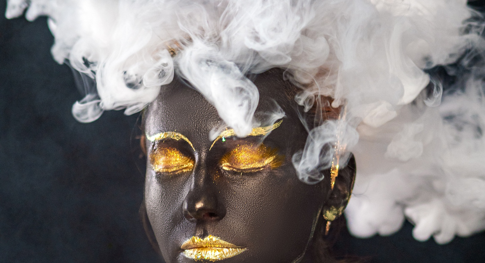 The head of a dark-skinned woman with gold makeup and heavy steam floating around her head instead of hair.